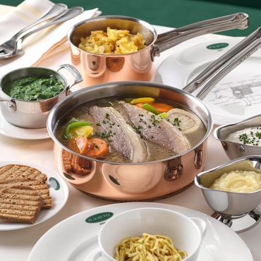 Ein Topf mit gekochtem Rindfleisch mit Gemüse, umgeben von Beilagen und Soßen auf einem weißen Tischtuch.