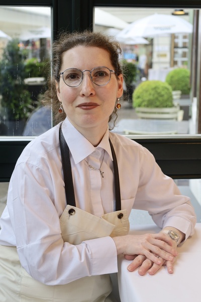 Frau mit Brille und Schürze sitzt an einem Tisch in einem hellen Café, die Hände gefaltet, und lächelt sanft in die Kamera.