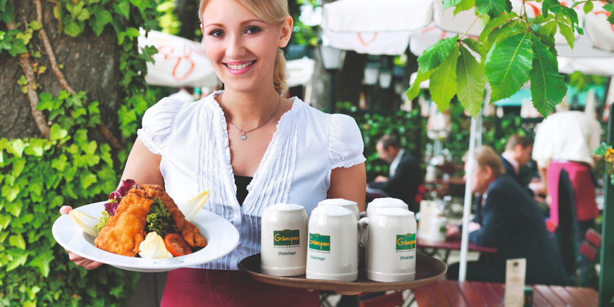 Eine lächelnde Kellnerin hält einen Teller mit Essen und ein Tablett mit Bieren in einem Restaurant im Freien.
